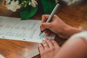 Une femme appose sa signature en bas d'un document sur un bureau en bois à côté d'un bouquet de fleurs blanches