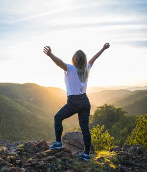 Une femme de dos qui arrive au sommet d'une montagne au coucher du soleil et qui lève les bras au ciel en signe de satisfaction